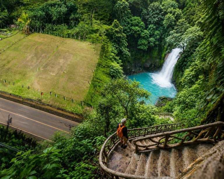 Person in red jacket standing on spiral stone staircase overlooking turquoise waterfall and lush green forest