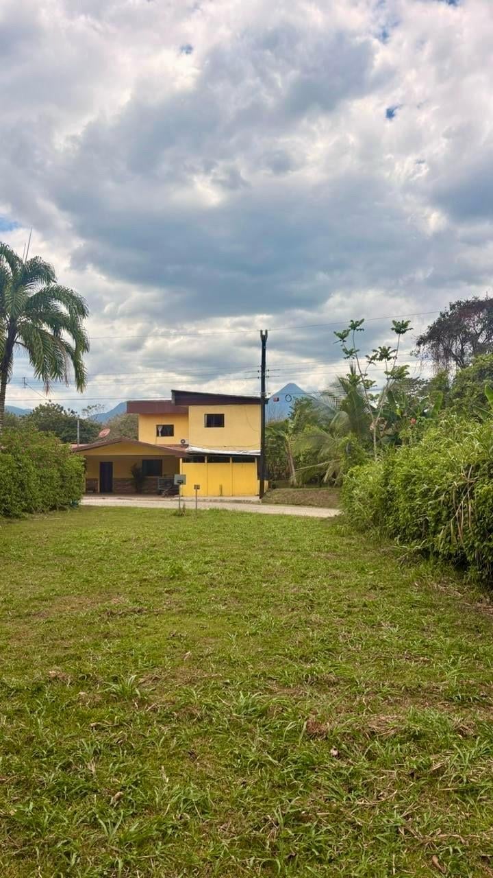 Yellow two-story house with flat roof on grassy property surrounded by tropical vegetation and mountains under cloudy sky