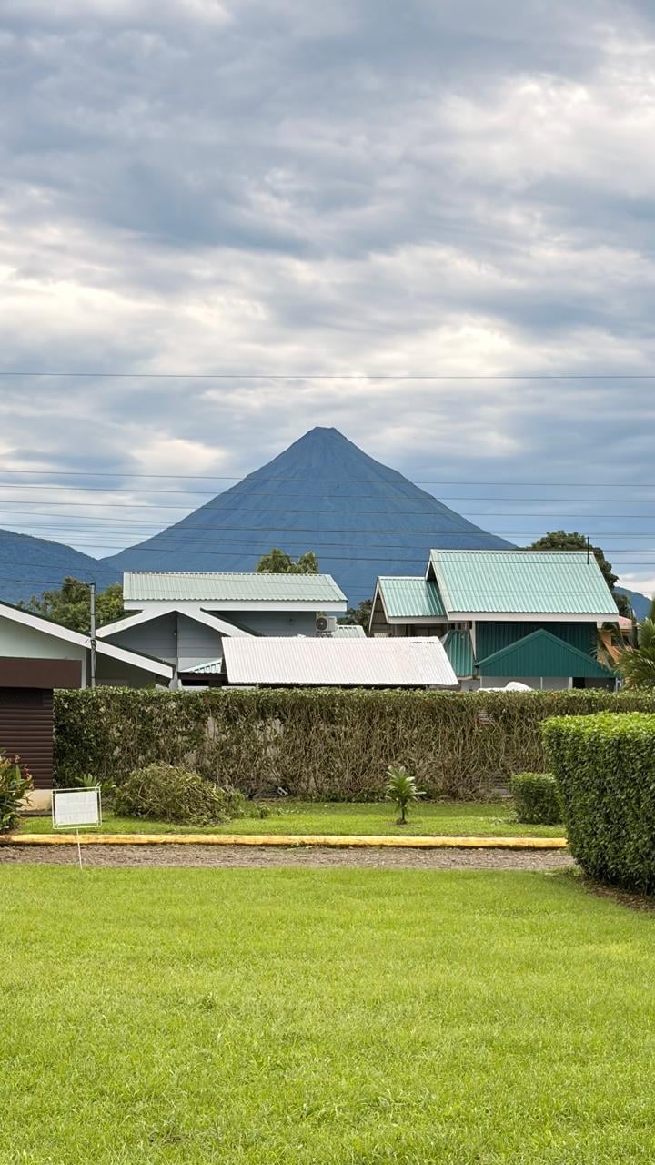 Green lawn and farm buildings with metal roofs overlooking a symmetrical cone-shaped mountain under cloudy skies
